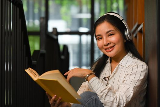 Smiling asian woman wearing headphone reading a book in library. People, knowledge and education concept.