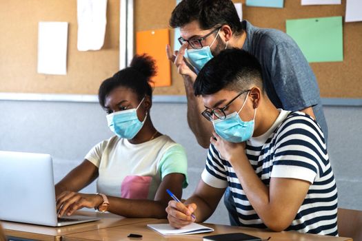 In class. Male teacher helps teen black girl and latino male high school students with lesson. Wearing protective face mask. Education concept.