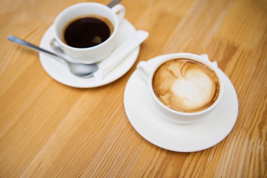 Top view. Close-up of white cups of aromatic coffee americano and frothy cappuccino on a wooden table, in the summer terrace of a cafeteria. Food and drink. Copy advertising space. Still life