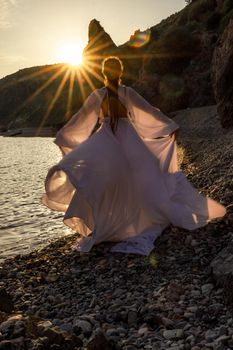 A mysterious female silhouette with long braids stands on the sea beach with mountain views, Sunset rays shine on a woman. Throws up a long white dress, a divine sunset