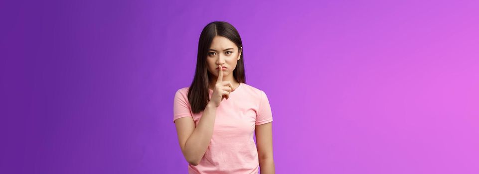 Shut up, seriously. Angry unsatisfied asian woman brunette frowning make hush sign, hold index finger on lips grimacing annoyed, prohibit speak, shushing stay silent, stand purple background.