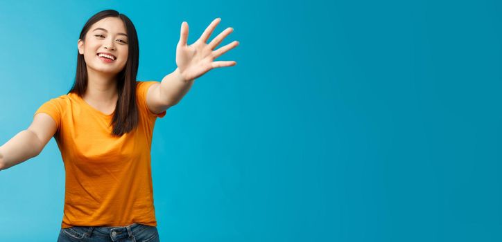 Friendly clingy girlfriend walking towards camera with extended hands to give tight hug, smiling broadly embracing friend, happily cuddling welcoming girlfriend, stand blue background.