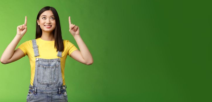 Dreamy hopeful cute asian girl look pointing up amused, smiling delighted, contemplate interesting object, enjoy stargazing, grinning satisfied, gazing top promo, stand green background.