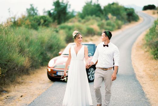 Bride and groom walk holding hands along the road against the background of the car. High quality photo