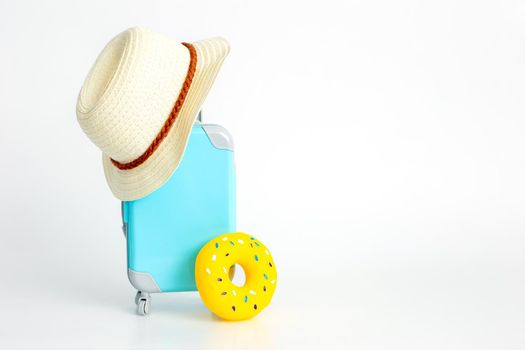 A blue plastic suitcase on wheels, a straw hat, and a yellow inflatable donut stand on a white background. copy space.