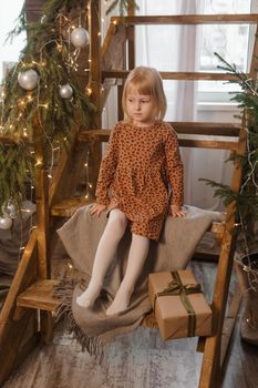 A little blonde girl is sitting on a wooden staircase in a Scandinavian interior decorated in a New Year's style. A child holds a Christmas gift in a craft package.