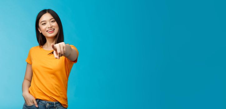 Lucky winner. Cheerful charismatic asian cute urban girl stand yellow t-shirt smiling friendly pointing finger camera choosing, picking person, inviting you team, stand happy blue background.