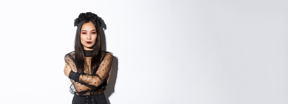 Young elegant woman in stylish gothic dress and wreath, embracing her body and looking sassy at camera, wearing halloween costume of undead widow, standing over white background.