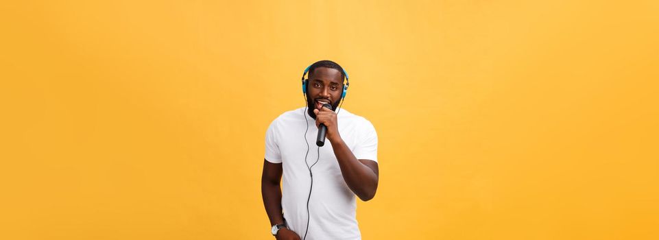 Portrait of cheerful positive chic. handsome african man holding microphone and having headphones on head listening music singing song enjoying weekend vacation isolated on yellow background.