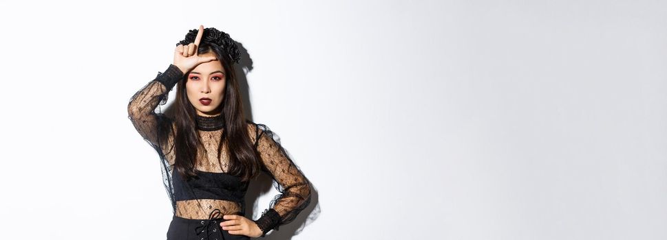 Image of arrogant sexy asian woman in witch costume showing loser gesture on forehead, mocking someone at halloween party, standing over white background.