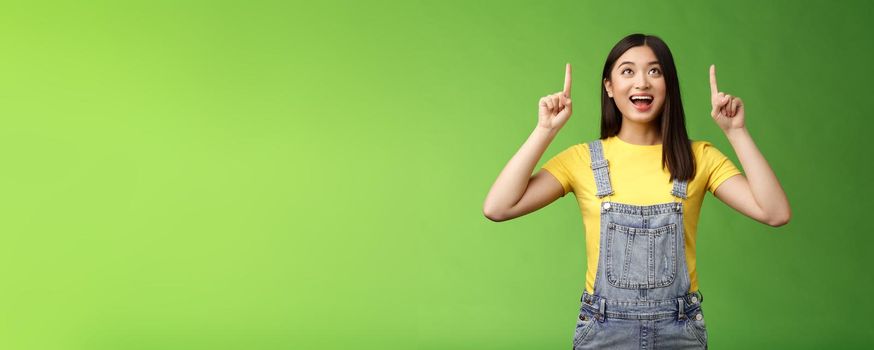 Carefree amused asian brunette in yellow t-shirt dungarees, smiling amazed and fascinated, look pointing up, observe cool new promo, grinning excited, enjoy watching top copy space.