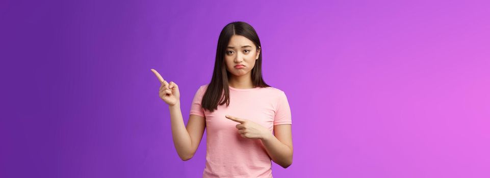 Upset unlucky cute sulking asian girl losing competition, pouting grimacing unhappy, pointing right, indicating fingers sideways with regret and sorrow, feel pitty herself, stand purple background.