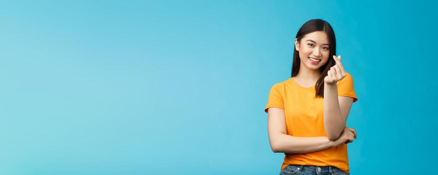 Cheerful cute asian woman dark short haircut, show korean heart sign with fingers, smiling joyfully, express sympathy and caring feelings, confess love stand blue background, grinning upbeat.