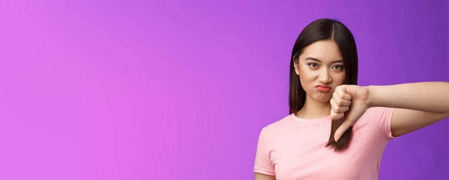 Close-up unsatisfied displeased judgemental asian girl give thumb down, grimacing, cringing from disappointment, not recommend awful product, complain bad service, stand purple background.