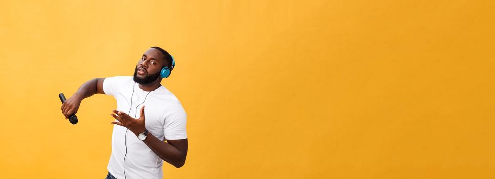 Portrait of cheerful positive chic. handsome african man holding microphone and having headphones on head listening music singing song enjoying weekend vacation isolated on yellow background.