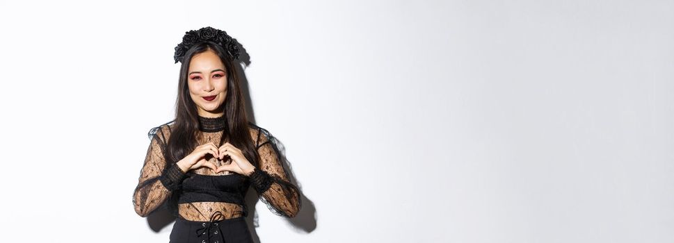 Image of lovely smiling asian woman love celebrating halloween, showing heart gesture and looking at camera, standing over white background in gothic lace dress with wreath.