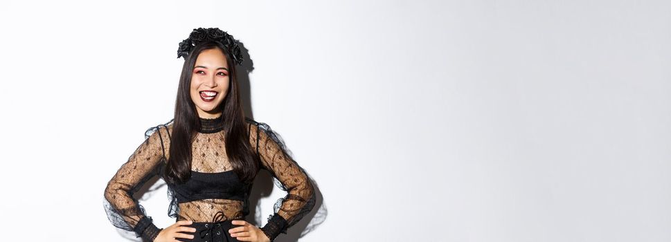 Image of carefree smiling asian woman in gothic dress and black wreath looking thoughtful at upper left corner, licking lips from desire or temptation, standing over white background.