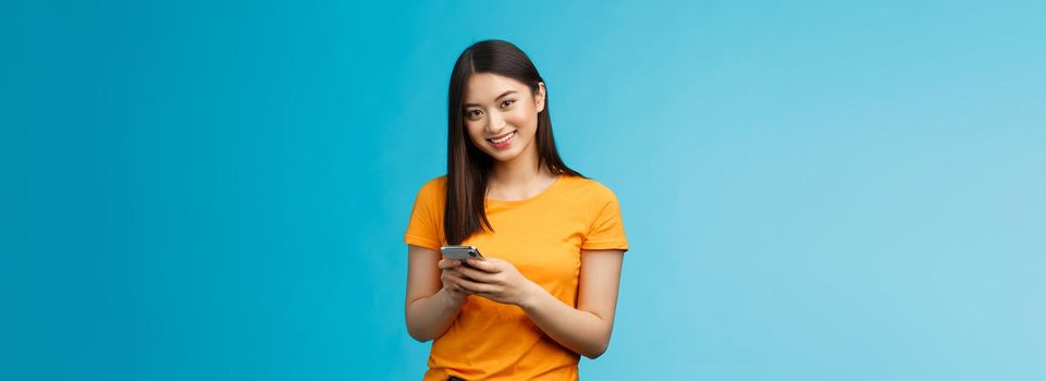 Cheerful friendly modern young asian girl student messaging friend, hold smartphone texting followers, using new internet application, try out app stand satisfied smiling camera, blue background.