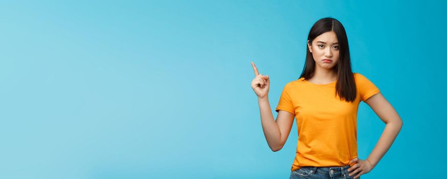 Moody upset asian girlfriend, sulking look offended and displeased pointing upper left corner, pouting disappointed, standing distressed blue background wear yellow t-shirt. Copy space