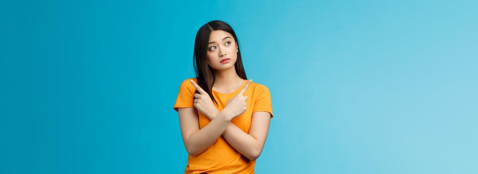 Confused cute asian female student thinking what do, look sideways thoughtful, cross hands chest pointing left and right, making choice, thinking what pick, taking important decision blue background.