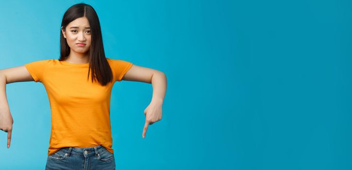 Moody sad asian brunette girl in yellow t-shirt smirking unsatisfied frowning bothered, complaining pointing down index fingers, showing bottom advertisement upset, jealous feel regret.