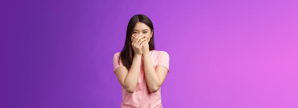 Cute carefree happy young asian girlfriend fooling around, giggle, close mouth palms not laugh loud, acting silly childish, joking, blushing receive surprising cute present, stand purple background.