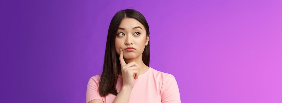 Close-up thoughtful cute asian girl dreamy standing purple background, touch cheek, turn left, thinking, doubting how act, wondering information, contemplating view from window.