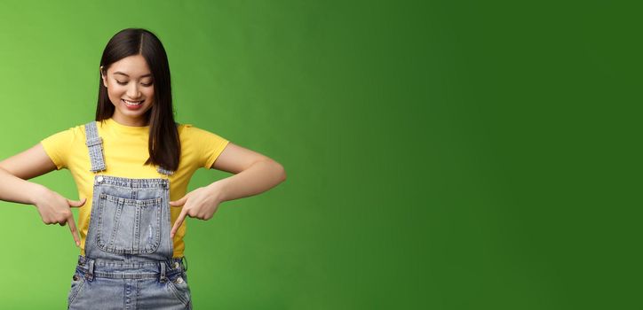 Studio shot easygoing cute asian female introduce new promo, pointing looking down amused pleasantly smiling, gladly show interesting thing, stand green background happy excited. Copy space