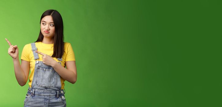 Upset cute asian female student pulling face regret shame, pity missing good chance buy product sale, pointing looking left disappointed, jealously gaze coworker, stand green background.