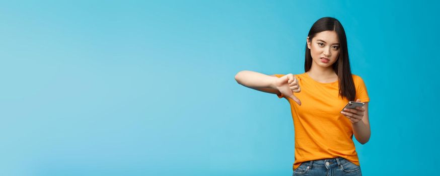 Displeased asian woman short dark haircut, frowning grimacing disappointed, show thumb down disapprove bad app, slow wifi speed hold smartphone unsatisfied, stand blue background. Technology concept