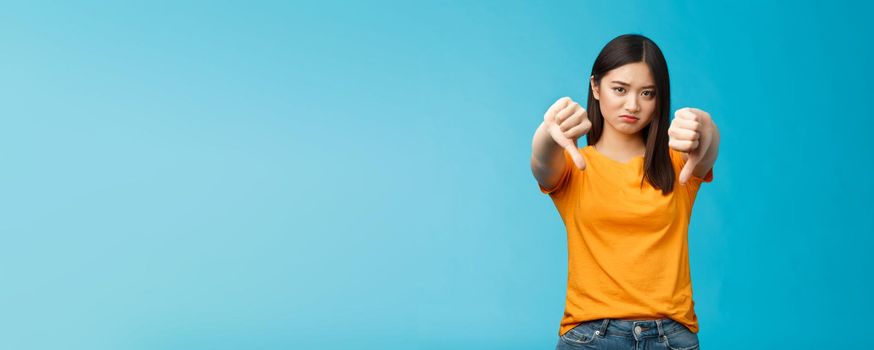 Upset gloomy asian girl with dark haircut frowning sulking sad, disappointed, show thumbs-down grimacing, give negative opinion, judging bad uninteresting movie, stand blue background.