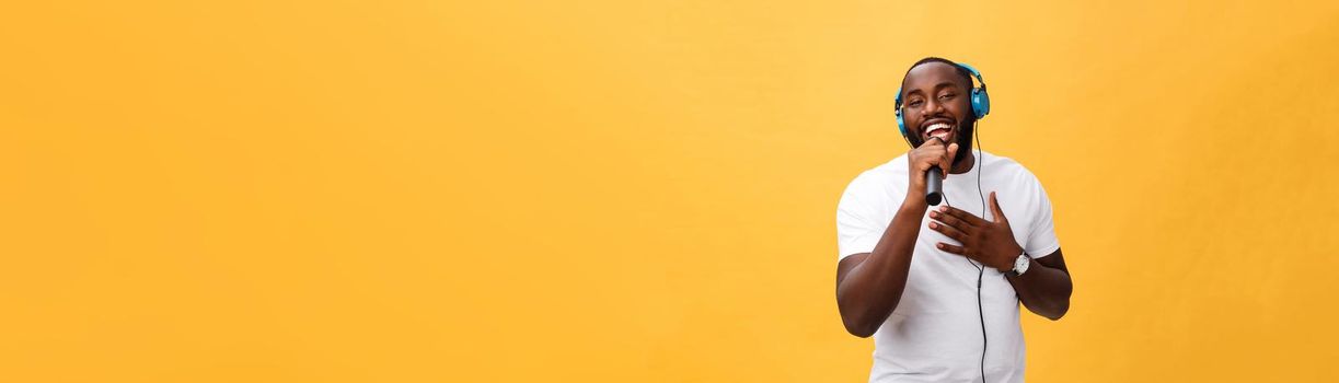 Portrait of cheerful positive chic. handsome african man holding microphone and having headphones on head listening music singing song enjoying weekend vacation isolated on yellow background.