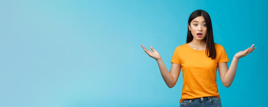 Questioned unaware young asian girl with dark short haircut shrugging hands spread sideways, look confused open mouth uncertain, being clueless, stand blue background wear yellow t-shirt.