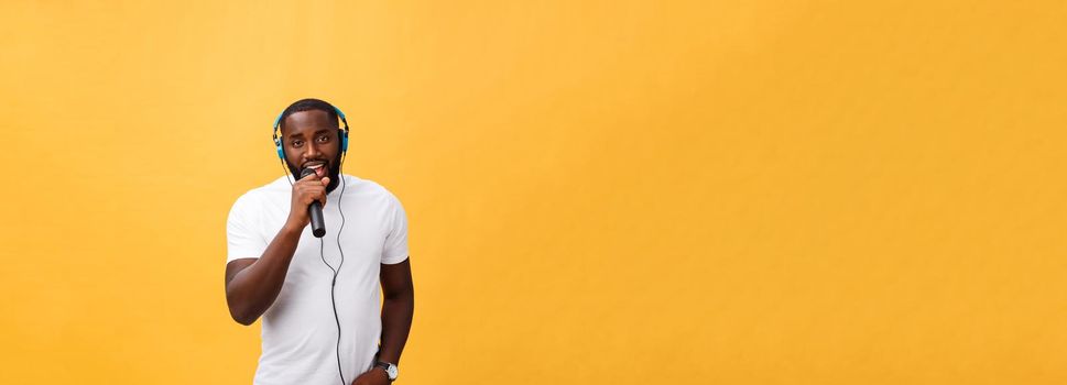 Portrait of cheerful positive chic. handsome african man holding microphone and having headphones on head listening music singing song enjoying weekend vacation isolated on yellow background.