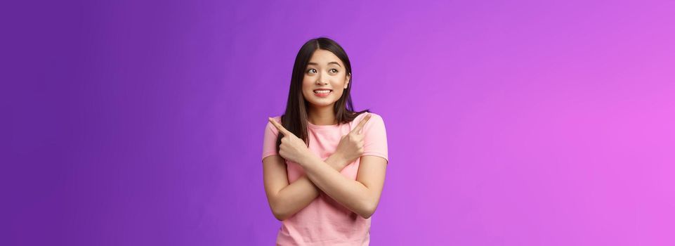 Cheerful timid cute asian girl, look tender, smiling gazing aside cross hands chest, pointing sideways, show left right, making decision, gathering confidence choose, purple background.