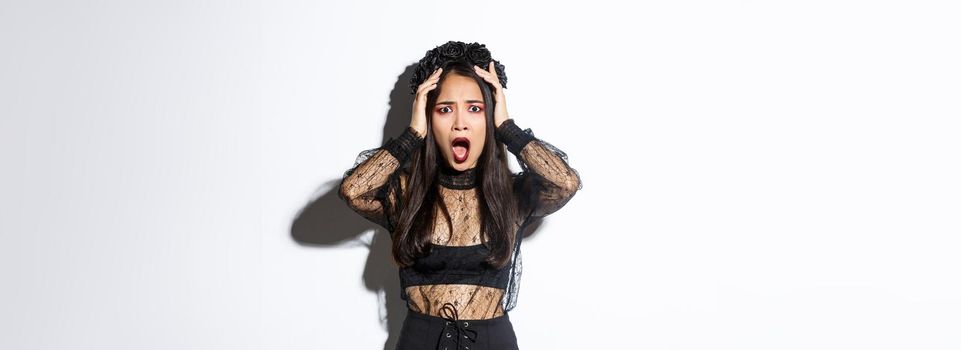 Image of horrified and shocked asian woman in gothic lace dress and wreath looking ambushed, wearing halloween costume, gasping and looking concerned, standing over white background.