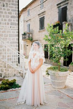 Bride in a lace dress with a veil stands in the courtyard of an old stone house. High quality photo