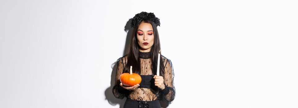 Myserious asian wicked witch in gothic dress, looking at lit candle, holding pumpkin, standing over white background.