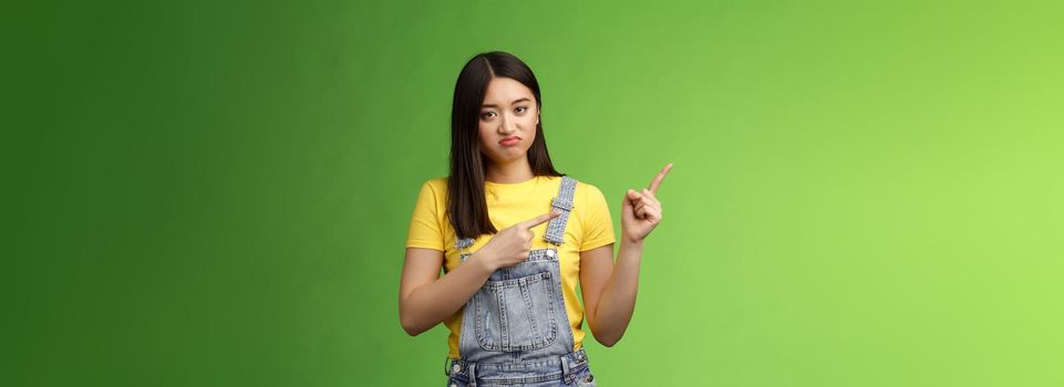 Disappointed uninterested asian gloomy girl pointing fingers left, grimacing sulking displeased, unwilling take part event, stare reluctant unimpressed, pose green background. Copy space