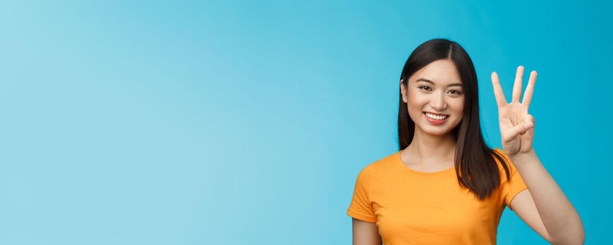 Friendly enthusiastic tender asian woman making reservation for three, number third, smiling broadly, stand blue background joyfully, posing near blue background in yellow t-shirt.