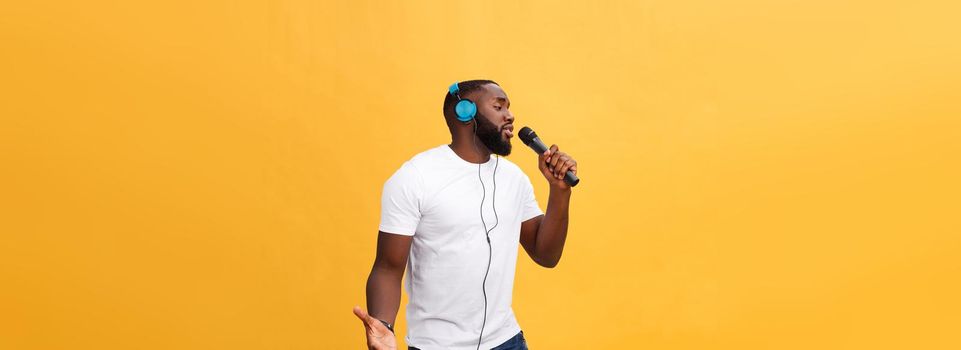 Portrait of cheerful positive chic. handsome african man holding microphone and having headphones on head listening music singing song enjoying weekend vacation isolated on yellow background.