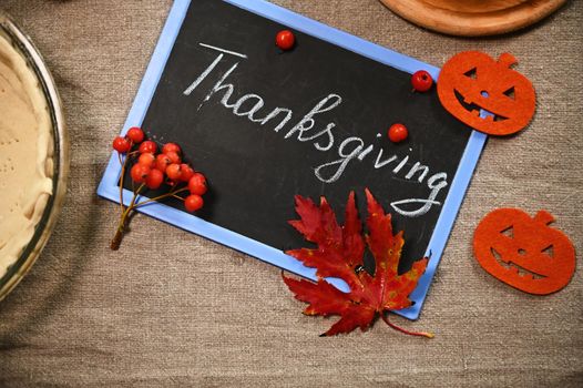 Thanksgiving written with a chalk on a blackboard, felt paper carved pumpkin, maple leaves and partial view of a rolled out dough on the baking dish, on linen tablecloth. Copy advertising space