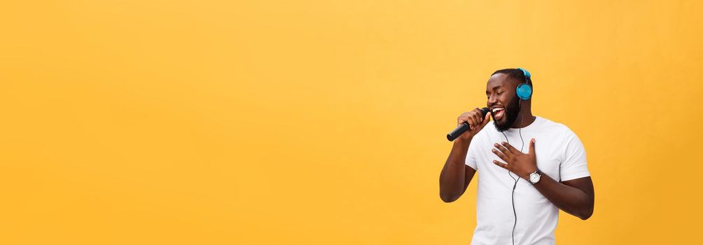 Portrait of cheerful positive chic. handsome african man holding microphone and having headphones on head listening music singing song enjoying weekend vacation isolated on yellow background.