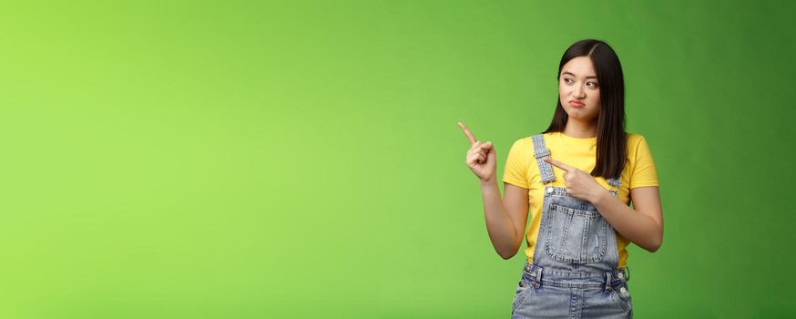 Upset cute asian female student pulling face regret shame, pity missing good chance buy product sale, pointing looking left disappointed, jealously gaze coworker, stand green background.