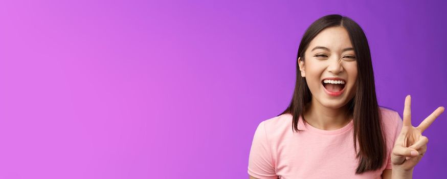 Headshot sincere carefree laughing asian girl having fun, show peace victory sign, joyfully look camera, express enthusiasm and happiness, enjoy funny friendly company, stand purple background.