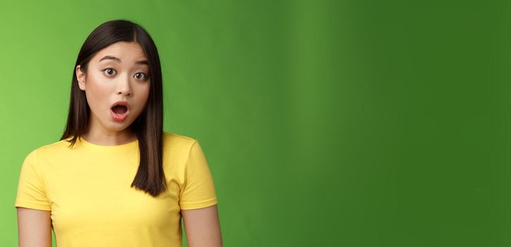 Close-up shocked amazed asian brunette female astonished hear stunning gossip, drop jaw stare camera speechless stand stupor green background, surprised what happening, wear yellow t-shirt.