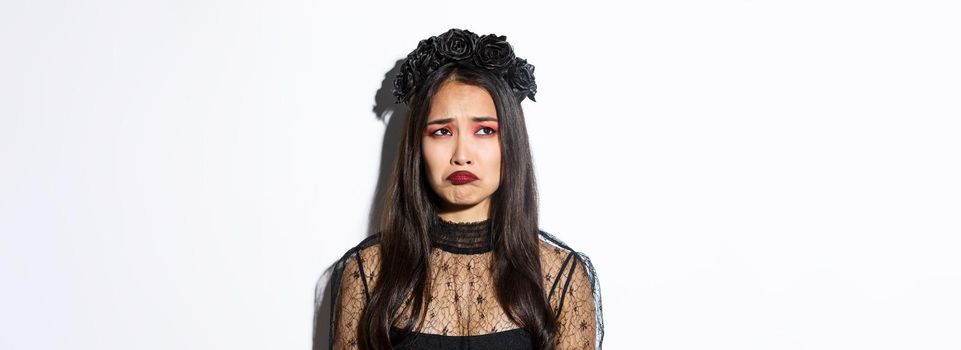 Close-up of sulking sad girl in witch costume looking at upper left corner disappointed, feeling miserable on halloween, standing over white background.