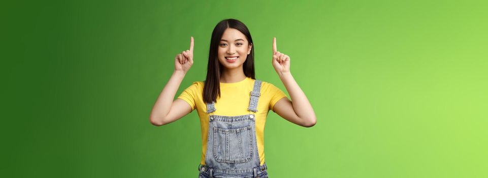 Friendly cute good-looking asian brunette female pointing up, raise fingers top advertisement, self-assured showing promo, smiling satisfied, introduce new product, green background.