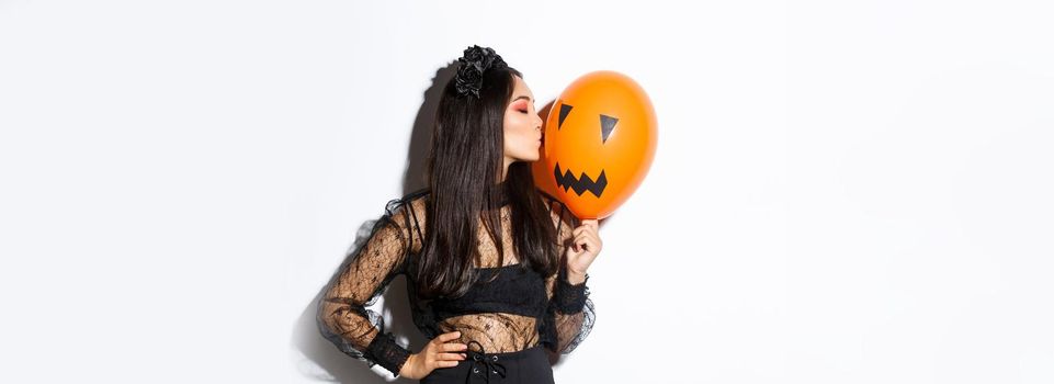 Stylish asian woman in gothic lace dress celebrating halloween, kissing orange balloon with face, standing over white background.