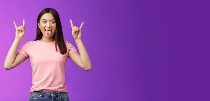 Rebellious amused asian girl enjoy rock-n-roll, attend awesome concert have fun, show tongue make heavy-metal gesture, express excitement and joy, stand purple background cheerful.